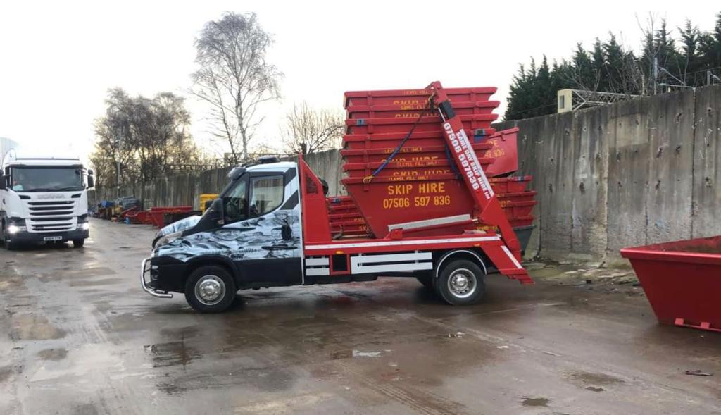 A small skip lorry carrying a red skip across a yard