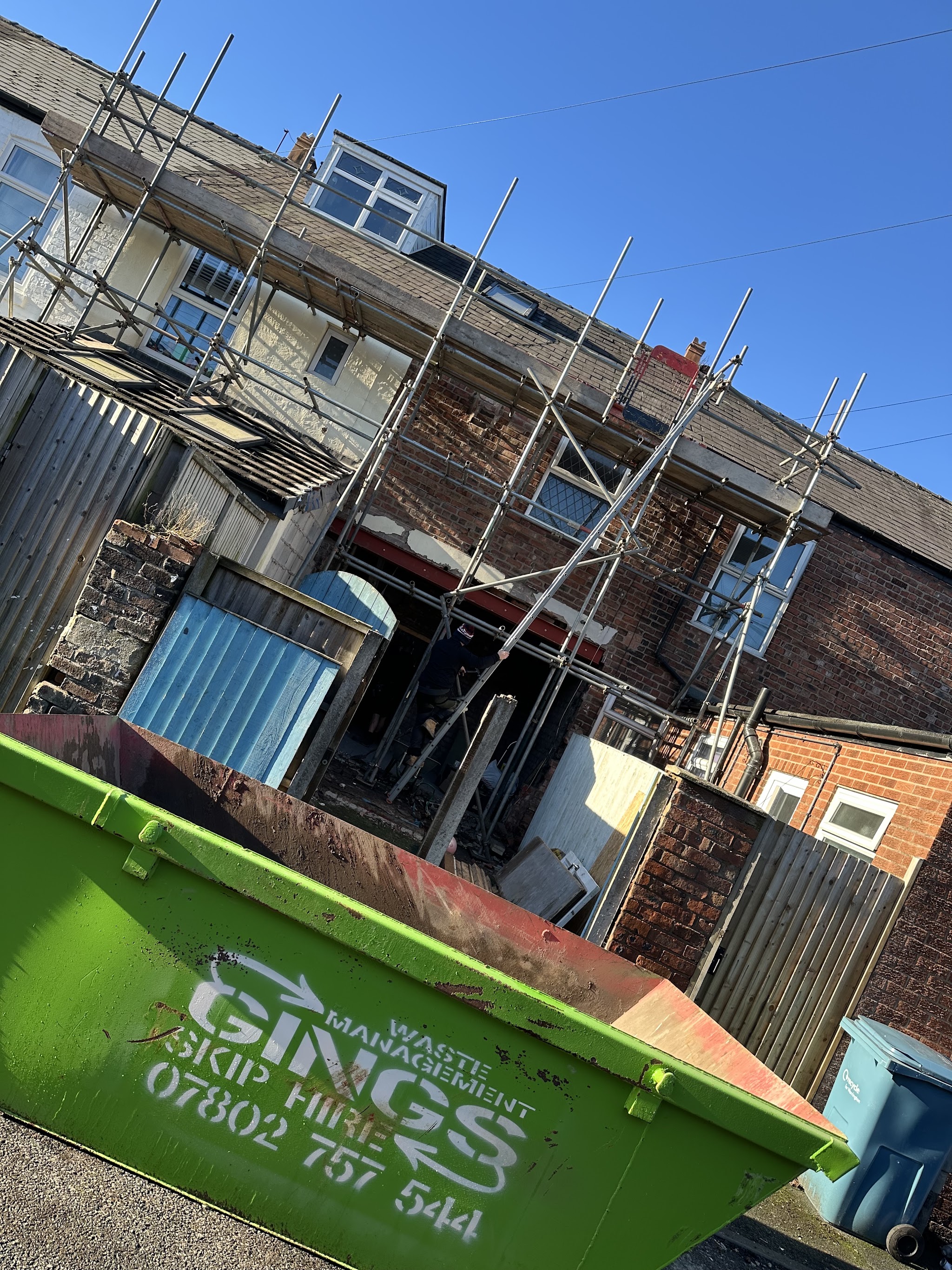 A green Gings skip beside a scaffolded brick house during building work
